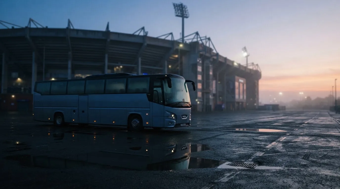 Autokar stoi na parkingu przed stadionem o świcie, rozmyte kontury trybun w tle, pusty plac i mokry asfalt.